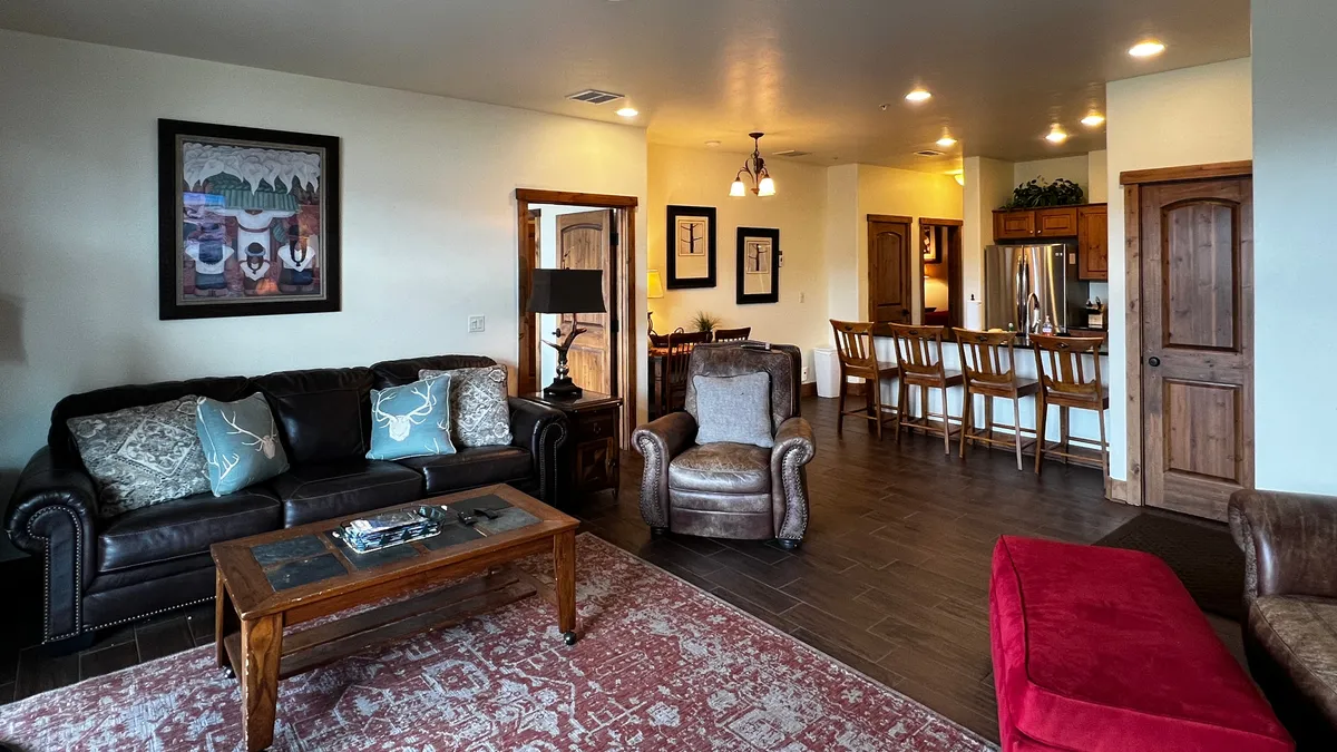 The image shows a cozy, well-lit area where the dining area and kitchen bar are visible in the background. The space is set up for socializing, dining, and entertaining, with a sofa and coffee table in the foreground and artwork to create a welcoming atmosphere.