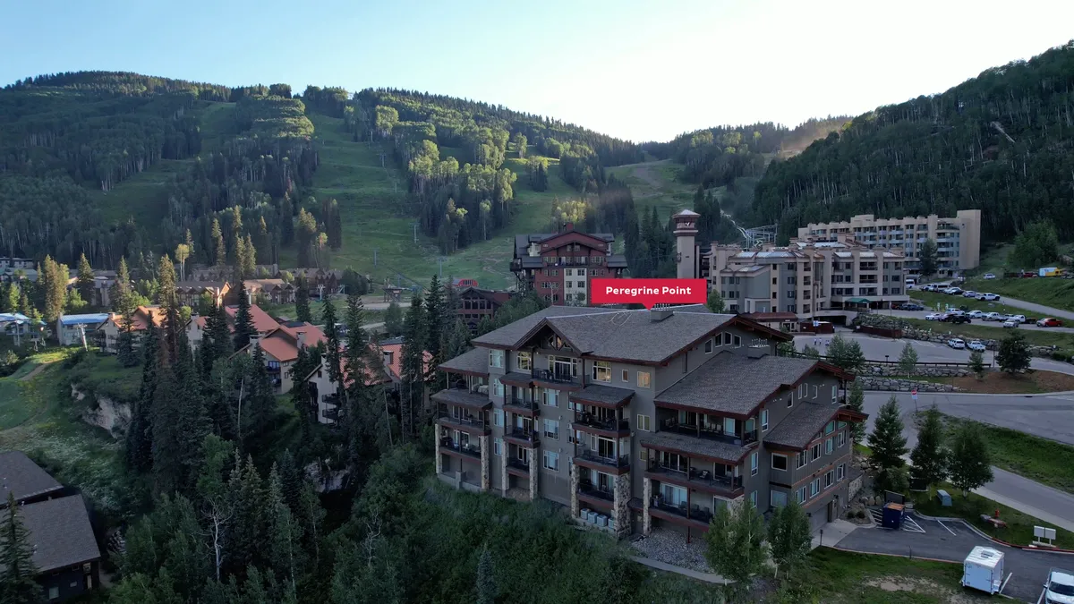 An exterior look at the Peregrine Point building, surrounded by mountains, with visible parking and garage access. The setting places the residence directly amid mountain scenery and hints at features like the heated garage and proximity to resort amenities.