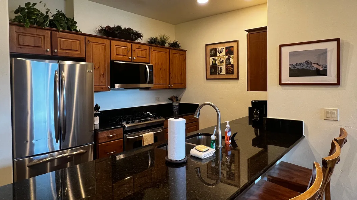The image showcases a warm, inviting kitchen with a sleek stainless steel refrigerator, a polished black granite countertop, and an island with comfortable seating—ideal for cooking, entertaining, and gathering. Framed wall art adds a touch of mountain-inspired decor, creating a cozy and stylish atmosphere perfect for memorable moments.