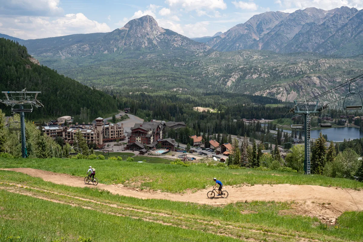 A panoramic view of mountain bikers navigating a scenic trail with the mountain landscape, resort area, and ski lift visible. The image transitions the narrative from winter sports to summer activities, emphasizing access to thrilling hiking and biking routes right outside the building.