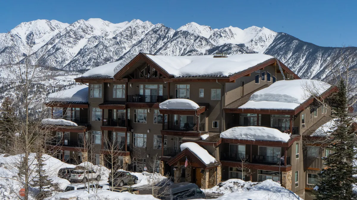An inviting exterior shot featuring a snow-covered roof, balconies, and the dramatic mountain range beyond. The tranquil winter landscape frames the condo as a cozy base for adventures and underscores the proximity to nature and the slopes.
