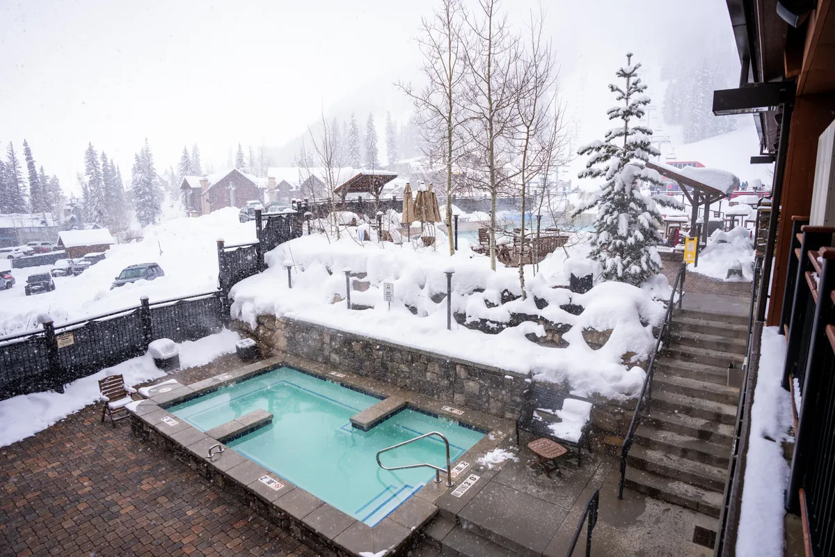 An inviting outdoor hot tub set amidst snow-covered pine trees and cozy cabins, with gentle steam rising into the cool air. Families and friends soak in the warmth, surrounded by a winter wonderland, while the sun sets over the snowy landscape.