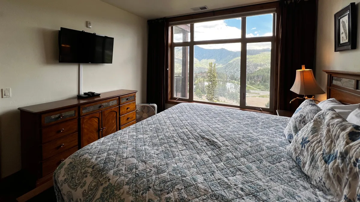 A tranquil bedroom with a large window framing breathtaking mountain views, a cozy bed with patterned bedding, and a wooden dresser topped with a TV. Soft natural light fills the room, highlighting the inviting atmosphere ideal for relaxation and private moments.