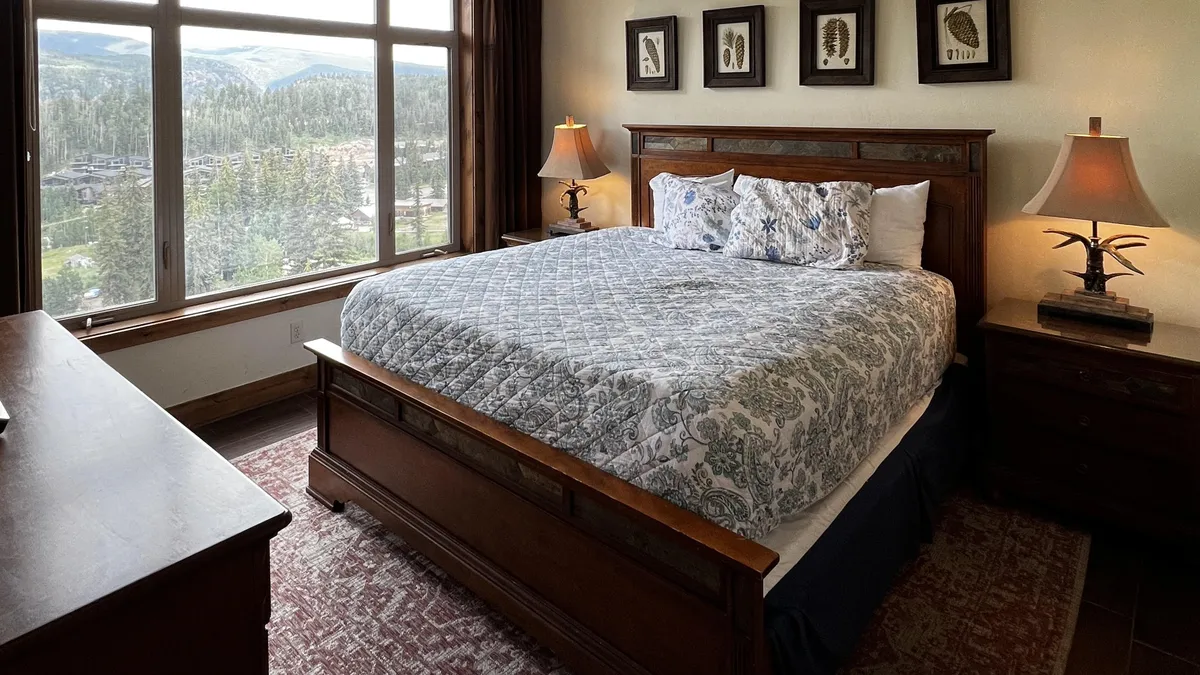 An inviting bedroom interior with a bed dressed in a patterned quilt, flanked by nightstands and lamps. A large window frames a stunning mountain view, allowing natural light to fill the space. The mood is tranquil and upscale, inviting relaxation after a day of adventure.