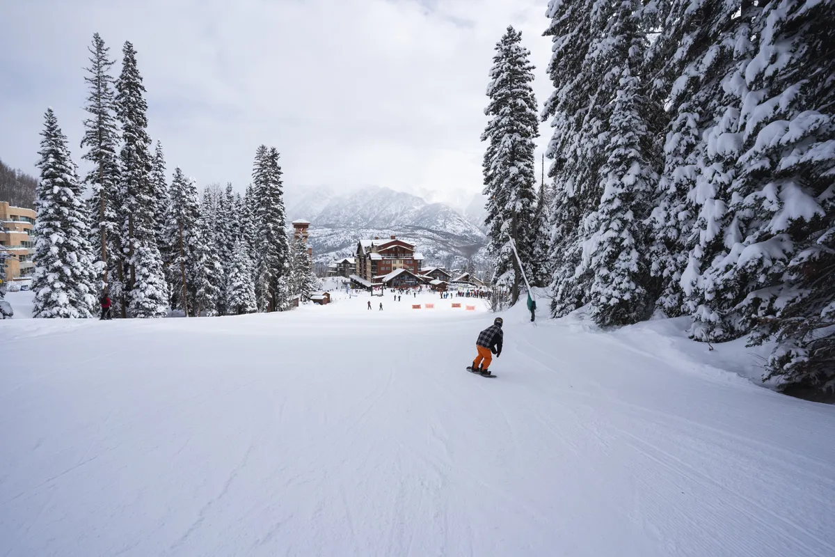 A lively, snow-covered slope with families and groups enjoying skiing and snowboarding. Pine trees border the white expanse, and a rustic ski lodge provides a cozy backdrop. The energy of people in action and the dramatic mountain scenery evoke excitement and togetherness.